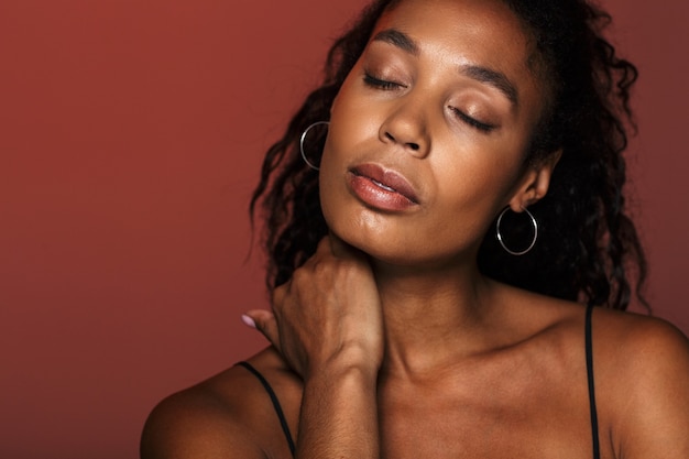 Picture of a beautiful pleased african young woman isolated over red wall with eyes closed touching neck.