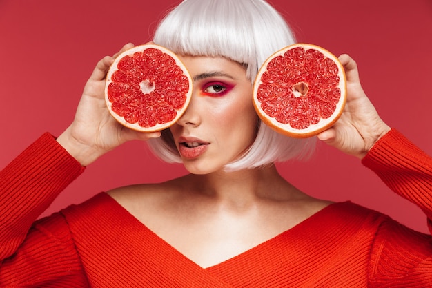 Picture of beautiful young woman isolated on red wall holding grapefruit.