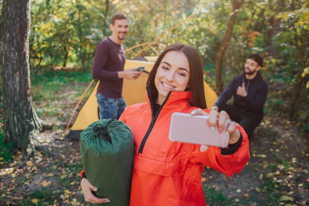 Изображение красивой молодой женщины принимает selfie и улыбки. Она держит спальный мешок. Молодые мужчины на спине тоже позируют. Они работают с палаткой.