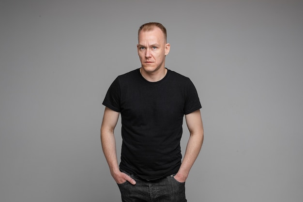 Picture of caucasian man keeps his hands in the pockets of his black jeans and looks to the left isolated on grey background