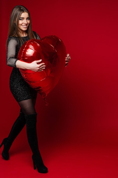 Picture of caucasian woman in black dress holds heart baloon in her hands on red