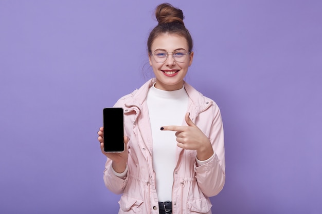 Picture of cheerful positive young female wearing stylish clothes, holding smartphone in one hand, showing direction with forefinger, smiling sincerely, presenting new device model. Tech concept.