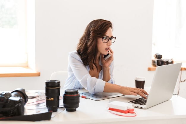 Picture of a concentrated serious young photographer woman in office working by laptop computer and graphic tablet talking by mobile phone.