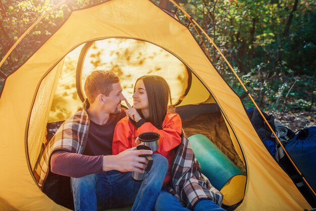 Picture of couple sitting together in tent. Young woman touches his nose with finger and smiles. He looks at her and hold thermos.