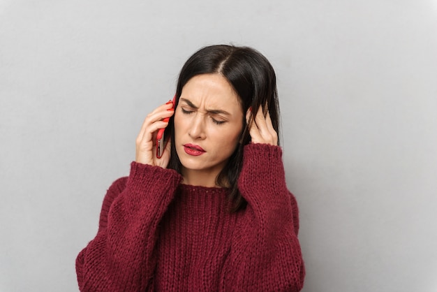 Picture of displeased young woman dressed in burgundy sweater talking by mobile phone isolated.