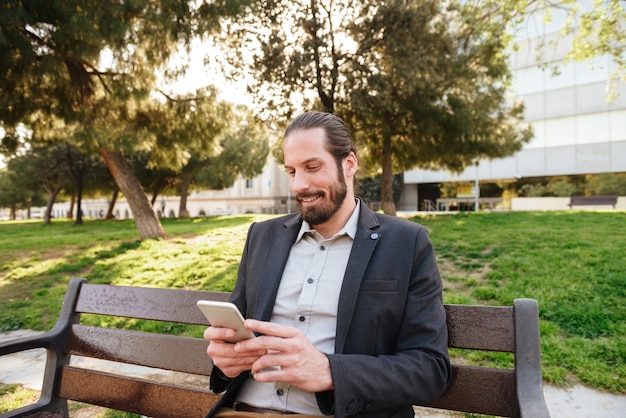 Picture of european bearded man 30s in formal wear holding and typing on cell phone, while resting in city park