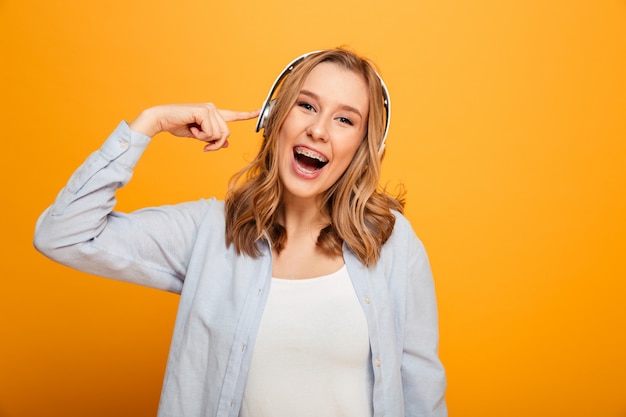 Picture of fascinating caucasian woman 20s wearing braces in casual clothing listening to music using wireless earphones, isolated over yellow space