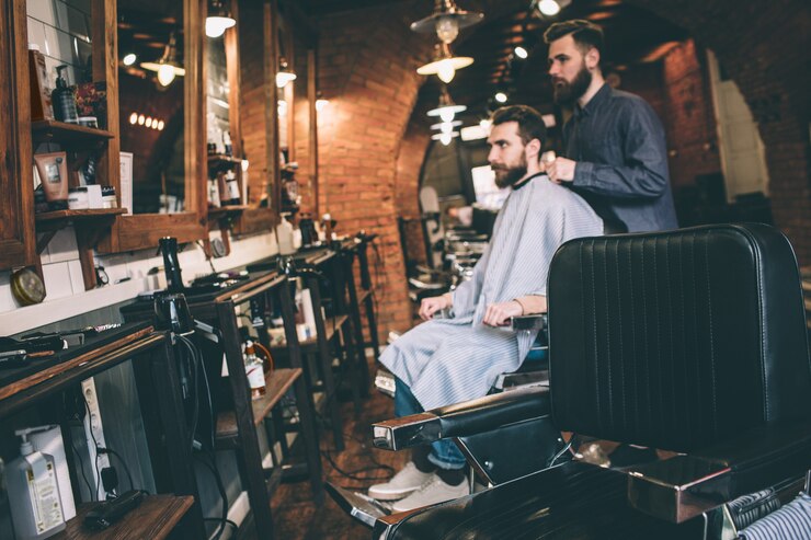 Photo a picture from a distance. guy is sitting in a chair and getting ready for cutting beard. his hairstylist is ready to start to work.
