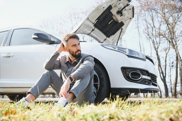 ボンネットが開いた壊れた車の隣に座っているイライラした男性の写真