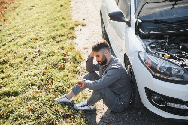 ボンネットが開いた壊れた車の隣に座っているイライラした男性の写真