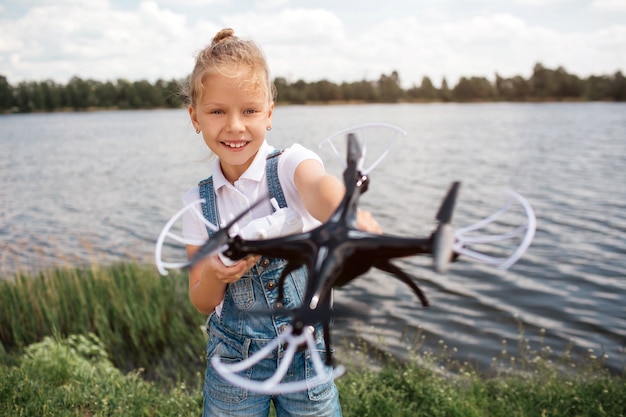 A picture of girl holding black drone i hands. She is looking at it. Girl is standing at the river shore.