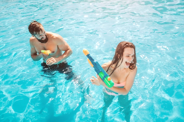 A picture of guy attacking girl in swimming pool. He is shooting from water gun. Girl is trying to defend herself and scream.