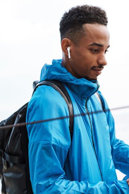 Picture of a handsome young african sports man outdoors at the beach sea walking with bag listening music with earphones.