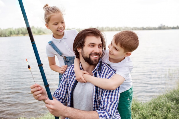 川岸で一緒に立っている幸せな家族の写真。男は彼を見ていると笑みを浮かべている間に少年はお父さんを抱いています。また、男は釣竿を持っています。女の子は父親の後ろに立って、彼を見ています。