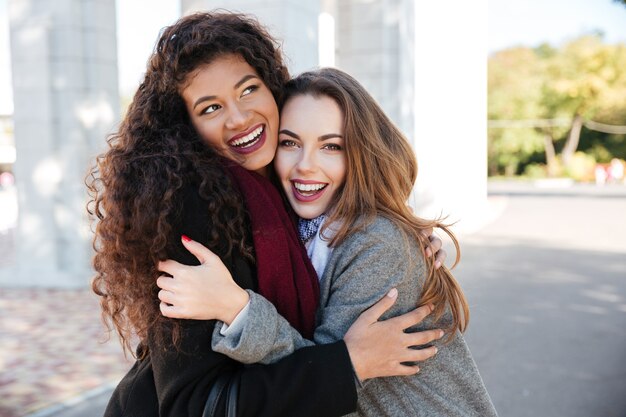 Photo picture of happy meeting of two friends hugging with street on background. caucasian girl looking at camera. african lady look aside.