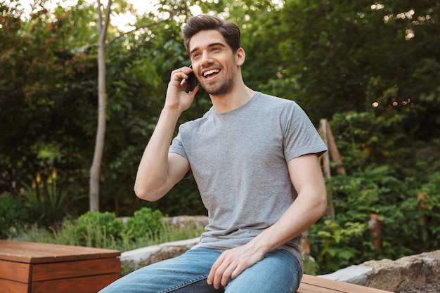 Picture of Happy young man in casual clothes talking by phone while sitting on bench outdoors