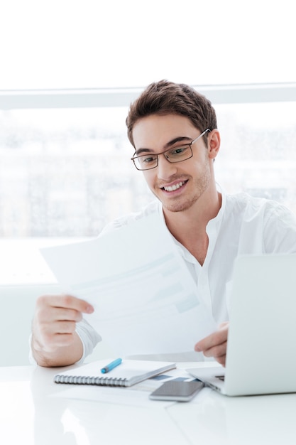 Picture of happy young man dressed in white shirt using laptop computer. Holding documents.