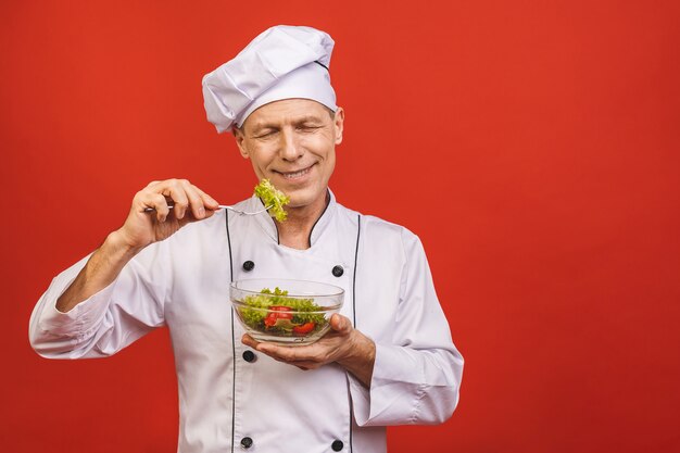 Picture of happy young senior chief cook in uniform standing isolated over red wall background, holding and eating salad.