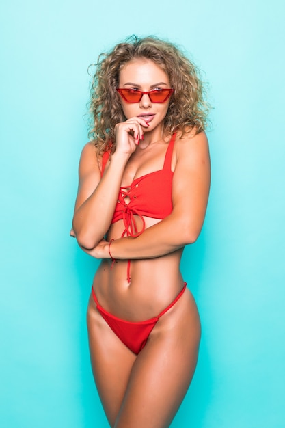 Picture of happy young woman in bikini and sunglasses with hands on chin isolated over blue wall.