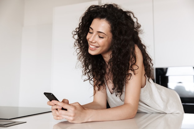 Picture of joyous lovely woman 20s with curly brown hair wearing silk leisure clothing smiling with perfect teeth, and using black cell phone while leaning on kitchen table