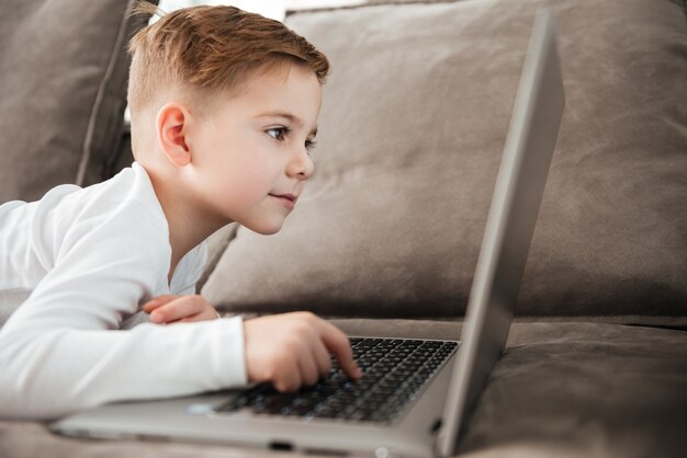 Picture of little cute boy using laptop computer while lies on sofa at home. Look at laptop.