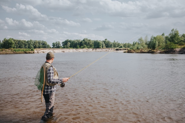 A picture of man standing in water and fishing. He is holding spinning in hands and looking at water. Guy has fishing net on the back. He looks calm and concentrated.