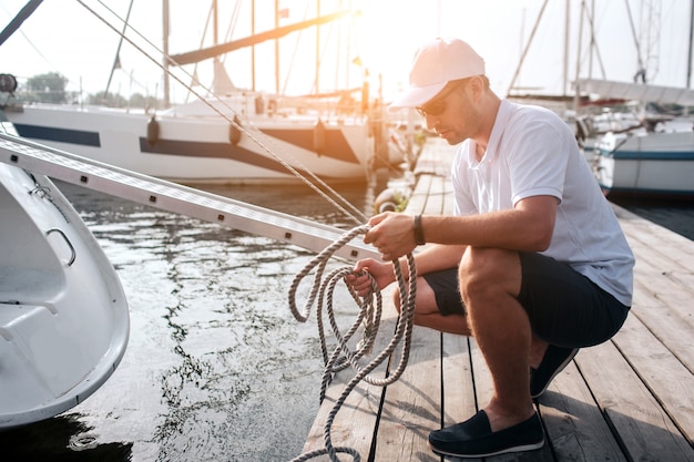 Picture of man in white cap and shirt sitting in squad position on pier. He holds lots of ropes. Guy is calm and peaceful. He is concentrated.