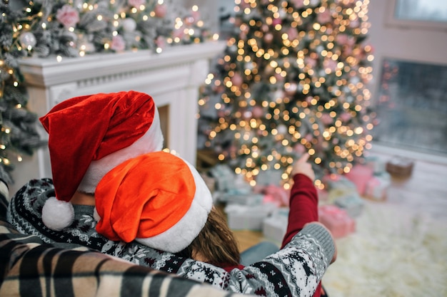 Picture of man and woman sit on sofa. They wear red Christmas hats. She points on Christmas tree. He embraces her. People are in decorated room.