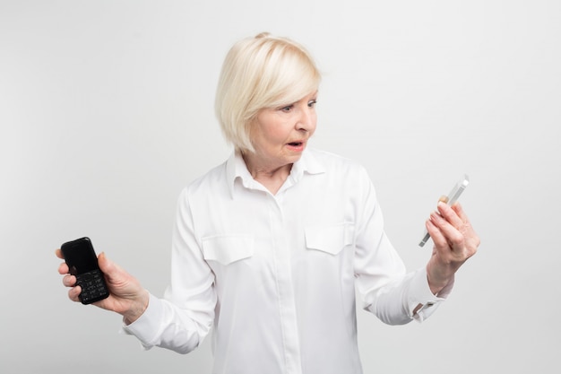 A picture of mature woman that holding two phones in her hands. She used to use the ld phone. The woman doesn't know how to use the new phone. It looks quite unusual.