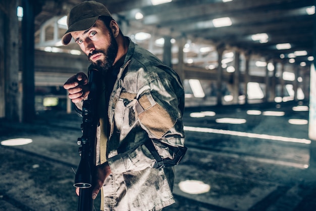 Un'immagine della posa del militare. è in un grande hangar. ha in mano un fucile nero e pronto a sparare. guy sta guardando dritto e mantenendo la calma. lui è molto silenzioso.