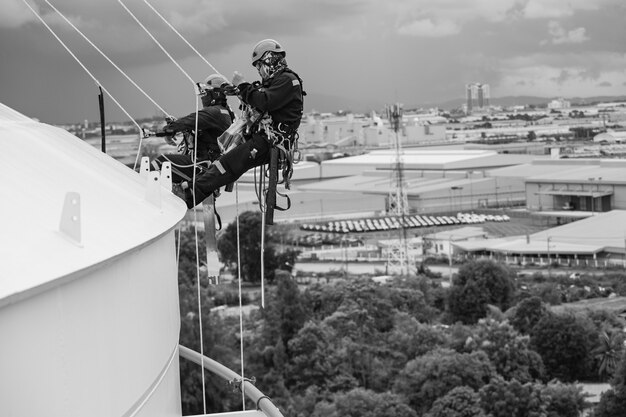 Picture monochrome male two workers control rope down top roof\
height tank rope access inspection of thickness shell plate storage\
tank gas safety work at height.