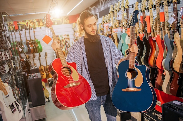 若いヒップスターの写真は、2つのカラフルなアコースティックギターを立てて保持します。赤と紺色です。男は2番目のものを見てください。男のエレキギターが彼の後ろにいます。