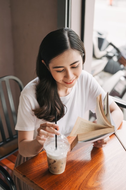カフェのテーブルに座って本を読んでいる若いきれいな女性の写真。