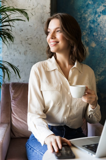 Picture of pretty beautiful young woman sitting in cafe indoors work with laptop using mobile phone.