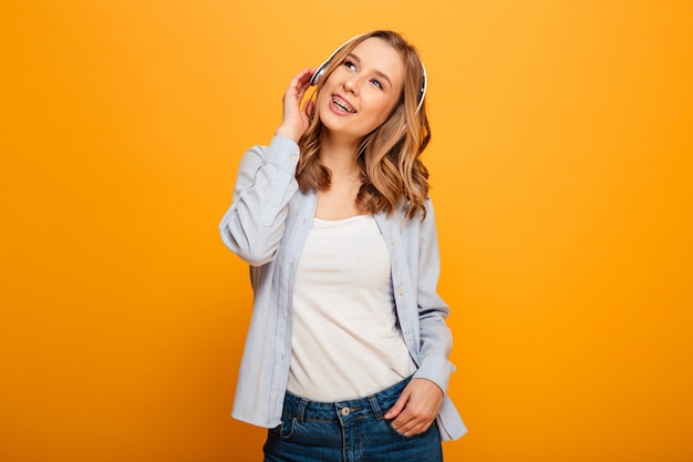 Picture of pretty caucasian woman wearing braces in casual clothing enjoying music via modern headphones, isolated over yellow space