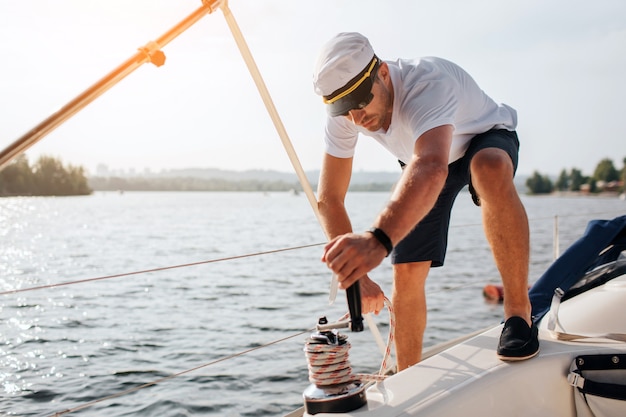 Picture of sailor stands on yacht and winds rope around. He is calm and concentrated. Young man works hard. He prepares for sailing.