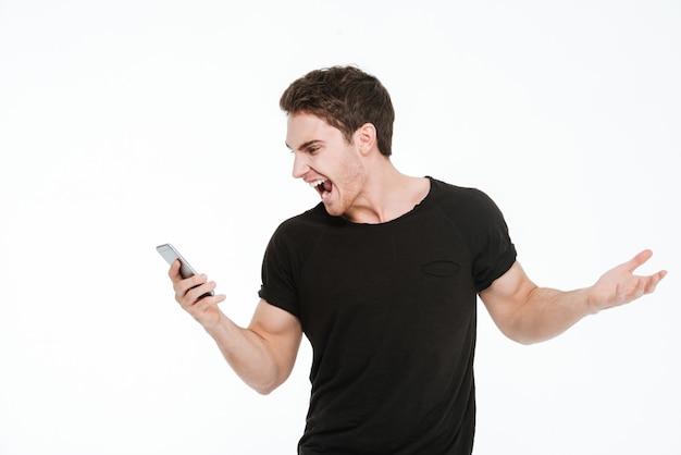 Picture of screaming young man dressed in black t-shirt standing over white background using phone.