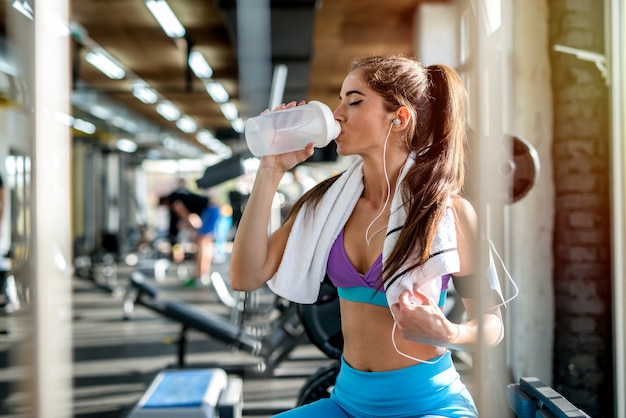 Picture of sexy fit girl sitting in a bight gym and drinking water. Listening to the music and resting after training,