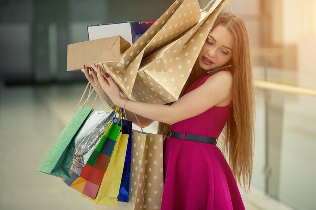 Picture of a shocked young brunette woman in white summer dress wearing sunglasses posing with shopping bags on pink.