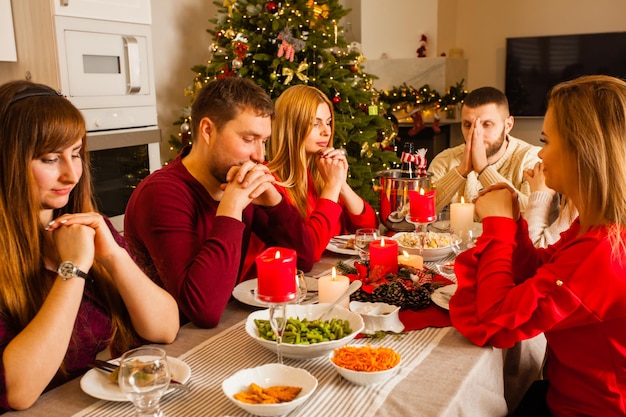 Picture showing group of young friends or adult sibblings with their couples praying around Christmas table. Winter holidays concept