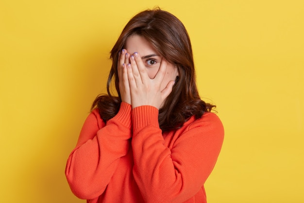 Picture of shy brunette teenage girl covering face with hands and peeping through her fingers . Young attractive female hiding her face being frightened, scared or ashamed, wears casually.