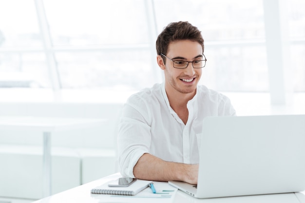 Picture of smiling young man dressed in white shirt using laptop computer. Look at laptop.