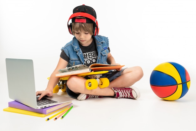 Picture of a teenager boy sits on floor in denim jacket and shorts. sneakers with yellow penny, red earphones, laptop, ball, and play computer games or do homework isolated