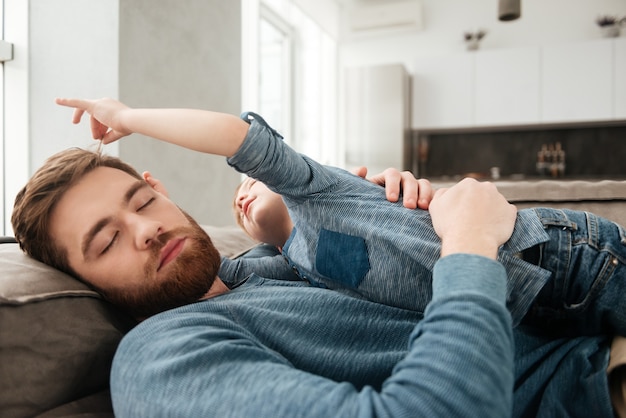 Immagine del padre barbuto stanco che dorme sul divano con il suo piccolo figlio carino.