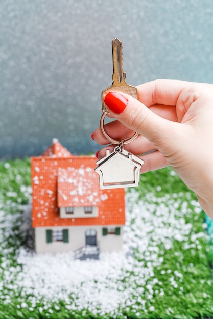 Picture of toy house with falling snow and hands with key on gray surface