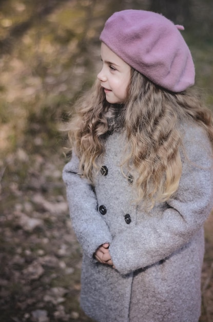 トレンディなビンテージ スタイルのベレー帽とコートで森の中を歩いている長い髪の少女の写真