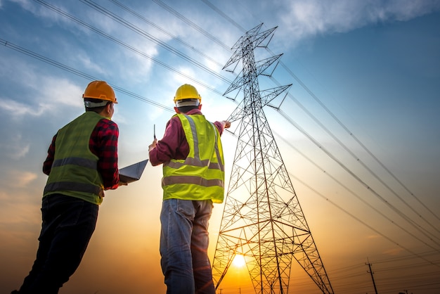 Фотография двух инженеров-электриков, проверяющих электромонтажные работы с помощью компьютера, стоящего на электростанции, чтобы увидеть работу по планированию на высоковольтных электродах.