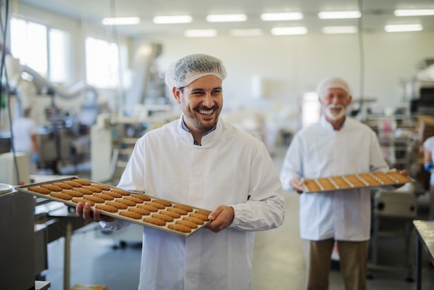 Picture of two happy smiling male employees in sterile clothes carrying trays full with fresh cookies in food factory. Helping each other and looking cheerful.