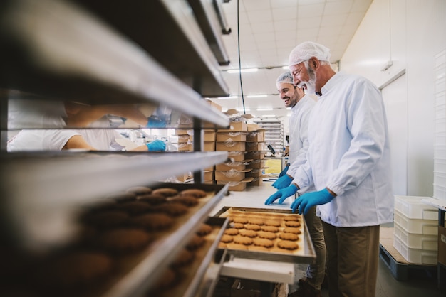 Picture of two male food factory employees in sterile clothes packing fresh made cookies. Standing in bright room.