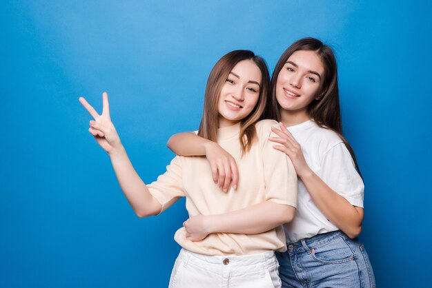 Photo picture of two playful girls standing together and showing peace gestures over blue wall